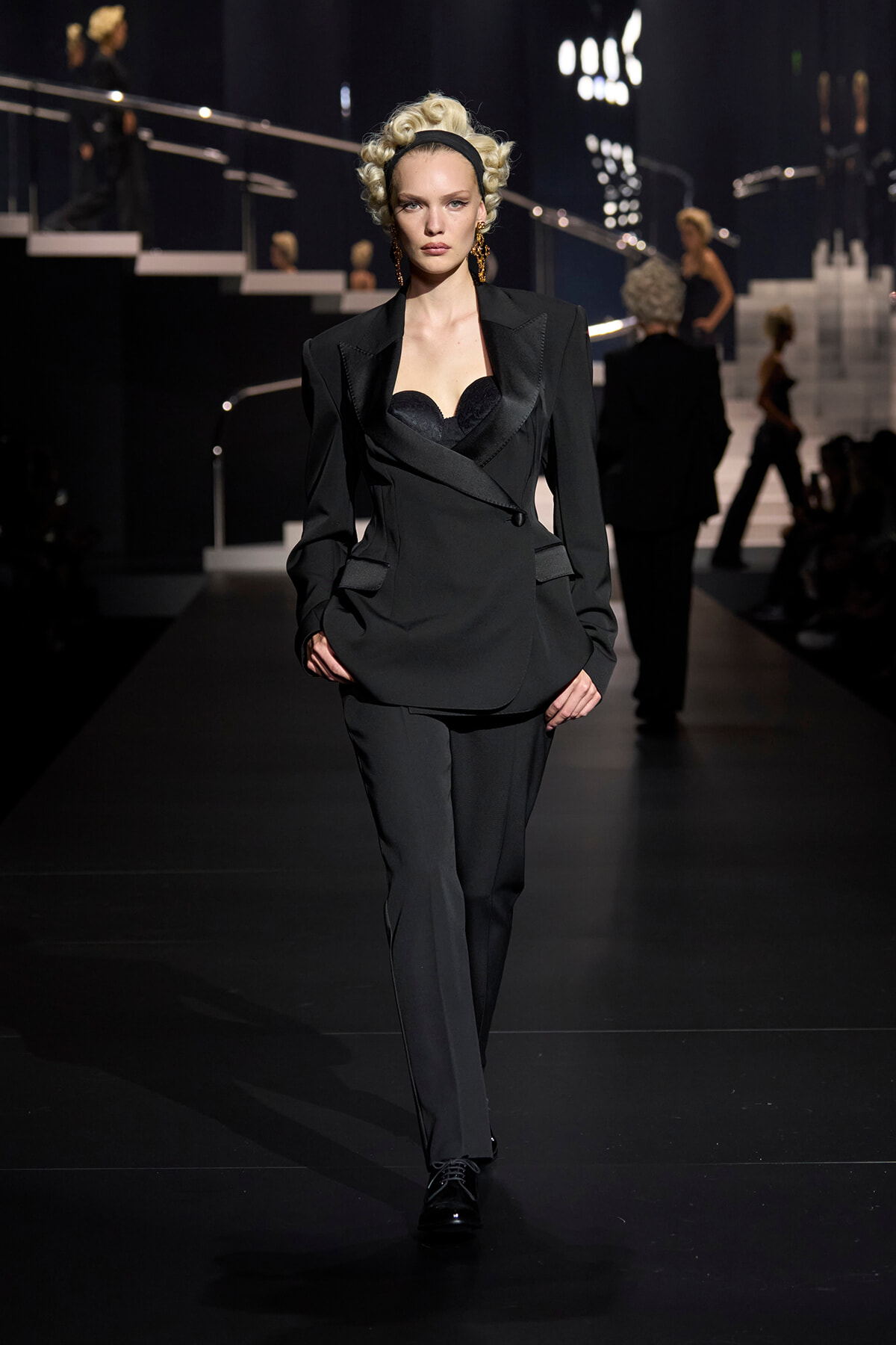 Runway model in a black tailored blazer and pants with a lace bustier, gold earrings, and vintage curled hair.