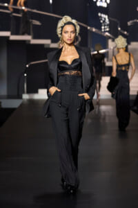 Model walking a runway in a black tuxedo-style outfit with a corset top and lace midsection, blonde curls, and oversized blazer, hands in pockets.