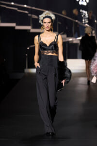 Model on runway in a black lingerie-style top with lace midsection, high-waisted corset belt, and wide-leg pants, holding a black fur stole.
