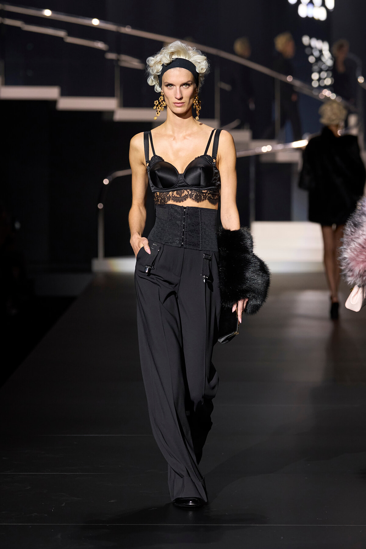 Model on runway in a black lingerie-style top with lace midsection, high-waisted corset belt, and wide-leg pants, holding a black fur stole.