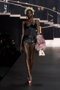 Model on a runway wearing a black corset top with a cinched waist, garter shorts, and a pink fur stole with a pale pink handbag. Rehearsal lighting highlights her dark skin and blonde curls.