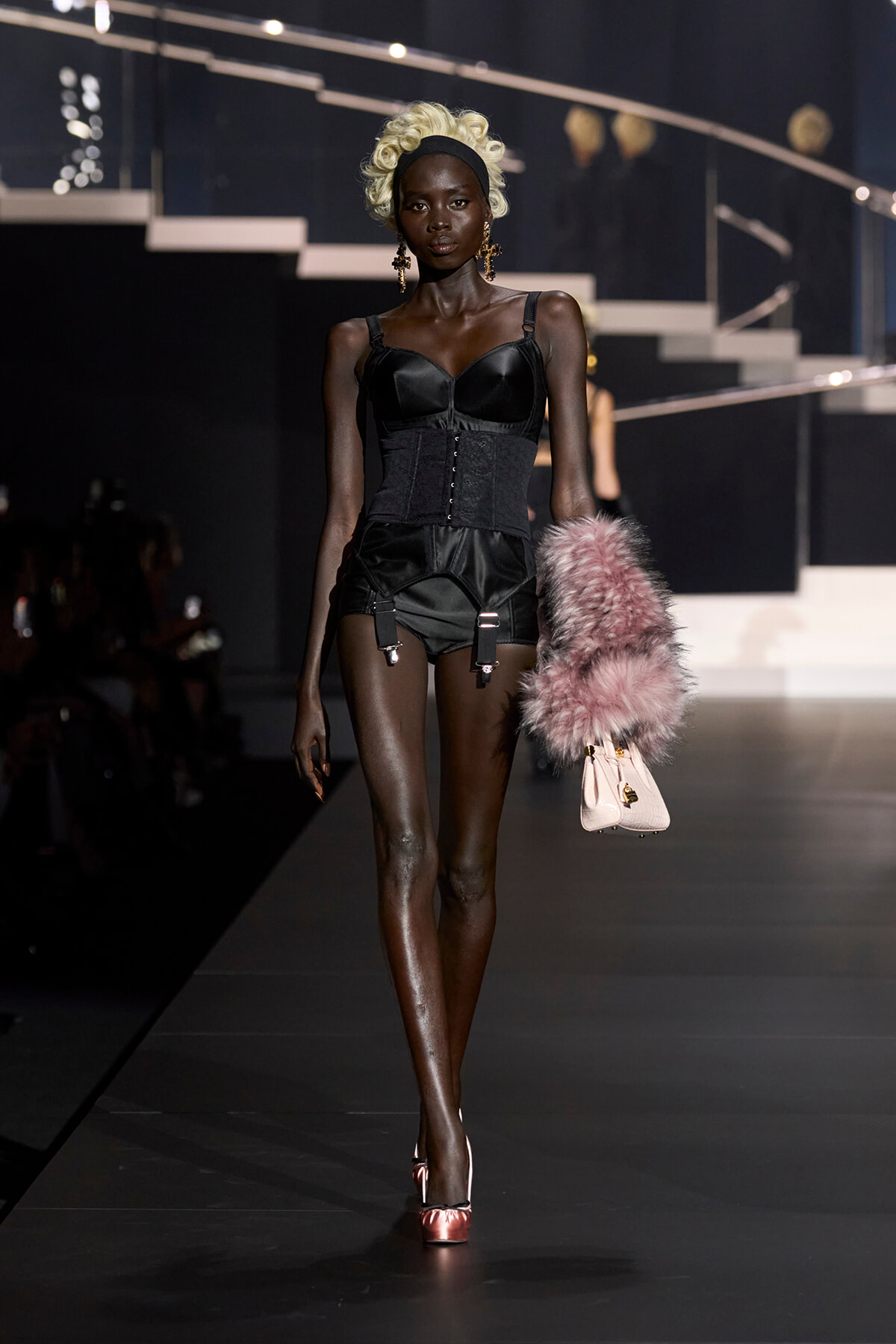 Model on a runway wearing a black corset top with a cinched waist, garter shorts, and a pink fur stole with a pale pink handbag. Rehearsal lighting highlights her dark skin and blonde curls.