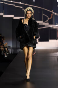 Model walking the runway in a long black fur coat over a black strapless outfit, with platinum blonde curls and a black headband.