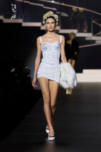 Model walks the runway in a white ruched mini dress with thin straps, holding a white fur stole and a small gold handbag.