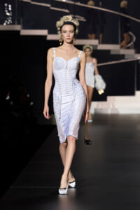 Model walks a black runway in a white lace-up corset dress with a headband and gold earrings, holding a small handbag.