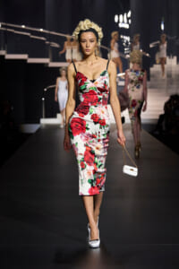 Model walks a runway in a white floral midi dress with red roses, holding a small white handbag with gold chain.