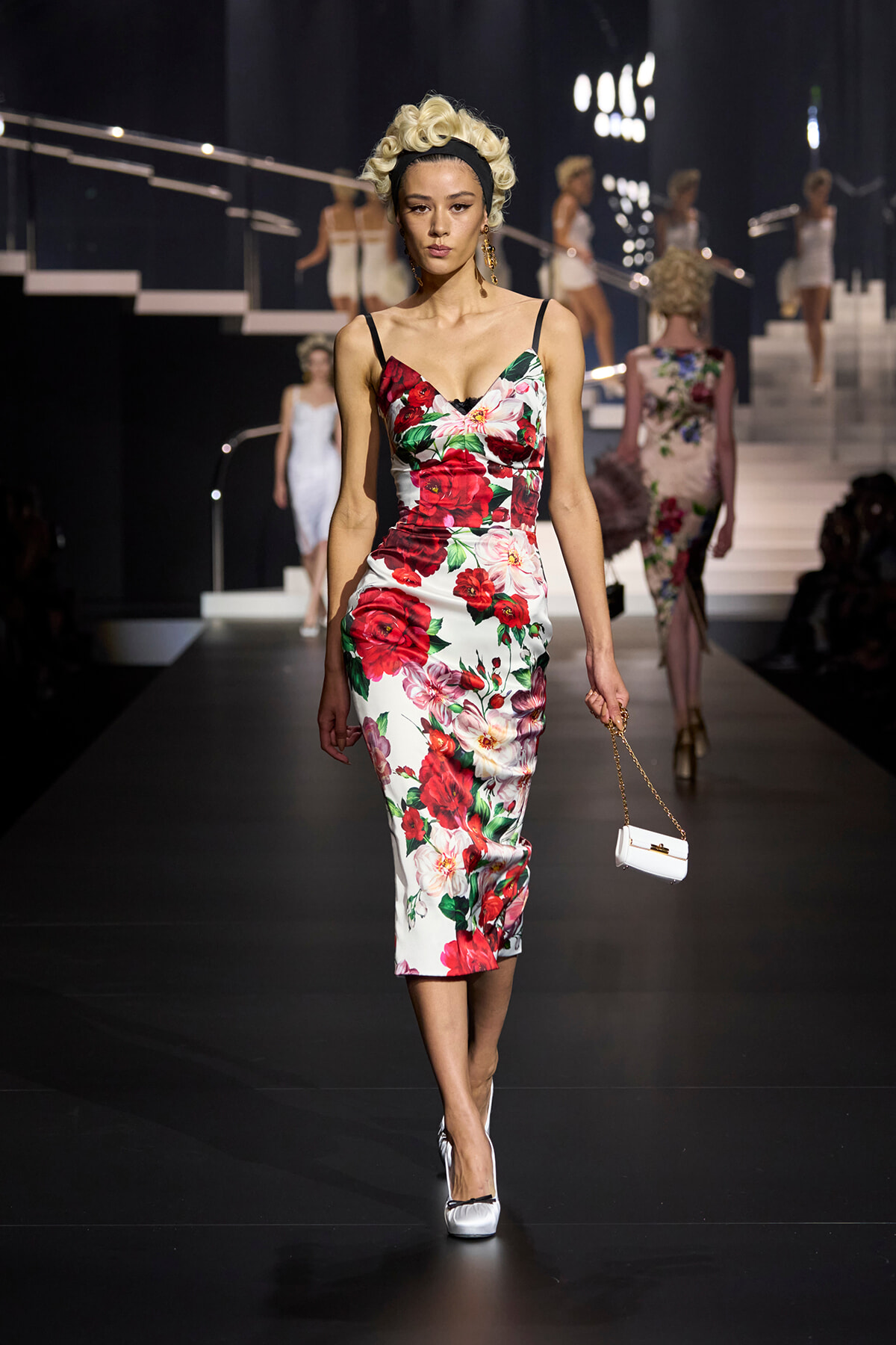Model walks a runway in a white floral midi dress with red roses, holding a small white handbag with gold chain.