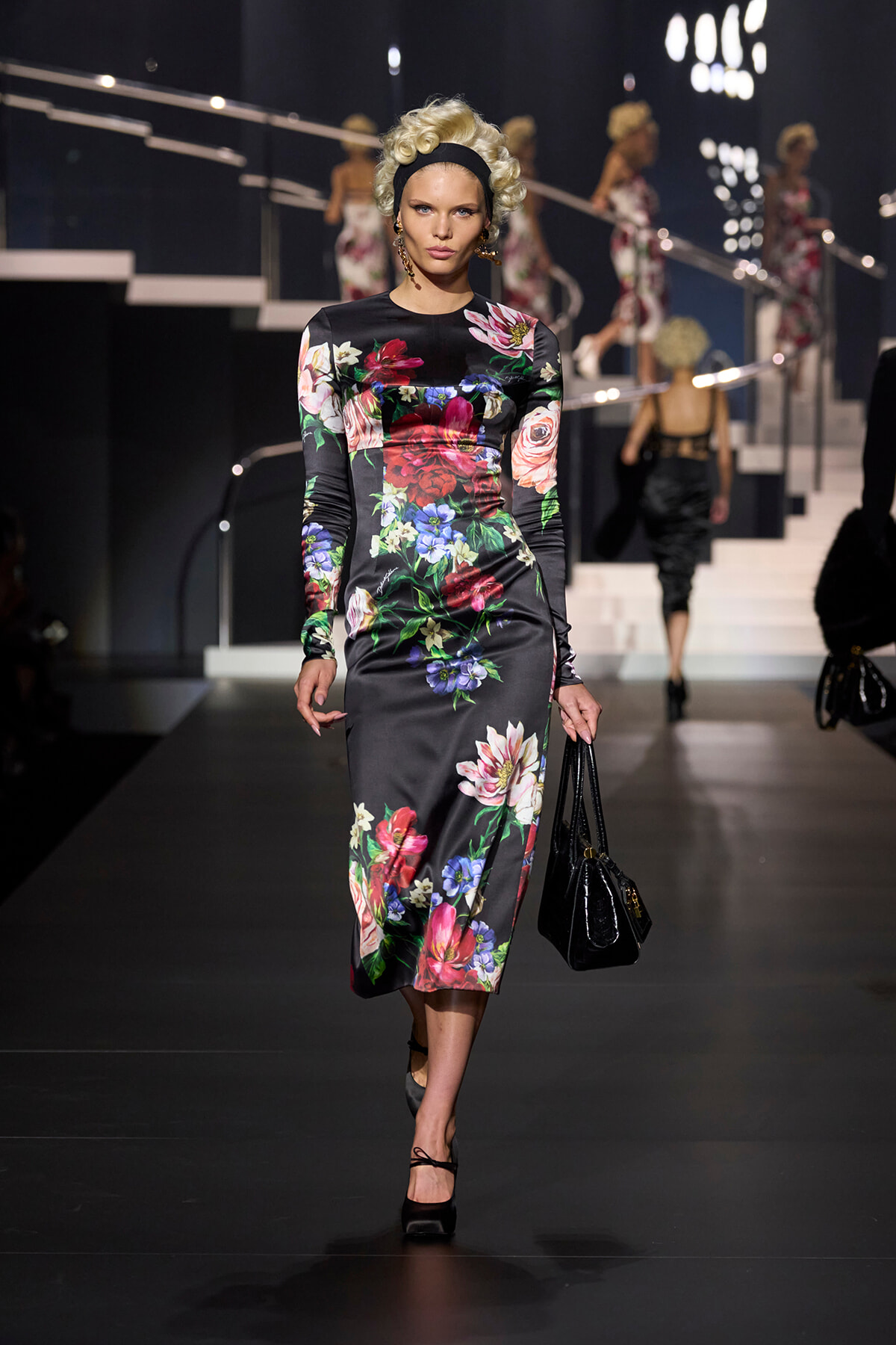Model walking a runway in a black satin floral midi dress, holding a black handbag.