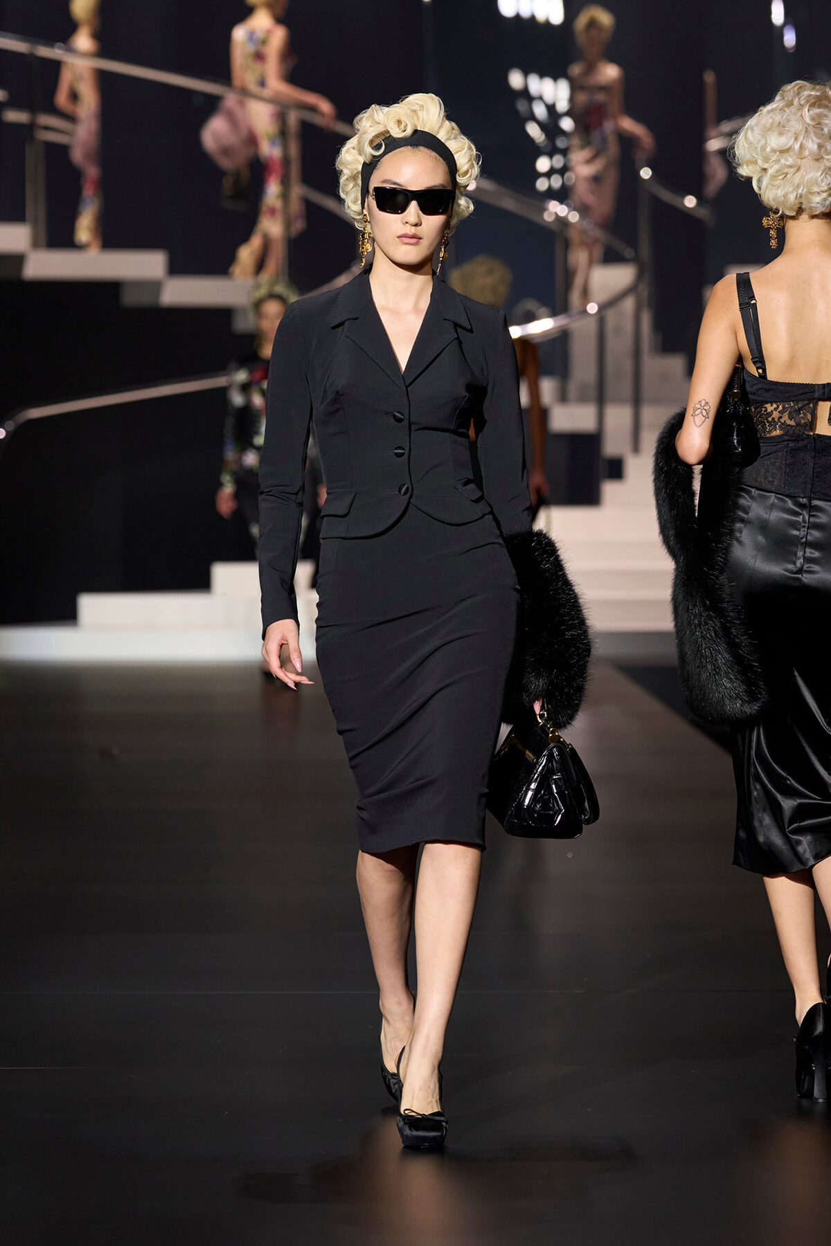 Model in a black tailored dress suit walking the runway; she wears oversized sunglasses, a blonde vintage hairstyle, and carries a black handbag.