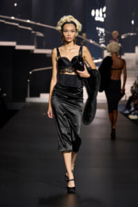 Model walking a runway in a black satin midi skirt and lace bustier, blonde curled hair, carrying a small bag and fur stole.
