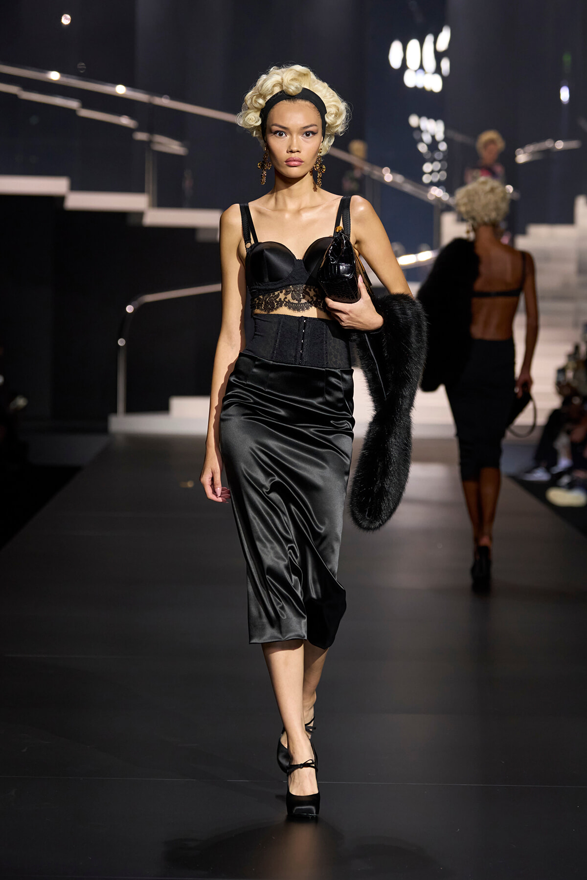 Model walking a runway in a black satin midi skirt and lace bustier, blonde curled hair, carrying a small bag and fur stole.