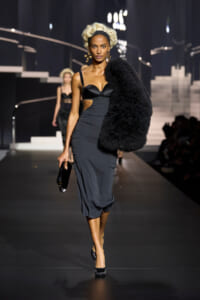 Model walks a fashion runway in a black midi dress with a fluffy black fur stole over one shoulder, holding a clutch and wearing black heels.