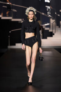 Female model walks a runway in a black cropped jacket and high-waisted shorts with pocket flaps, carrying a small black handbag with a gold chain.