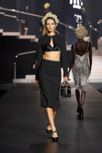 Runway model in a black cropped jacket, bra-style top and midi skirt, carrying a small black handbag.