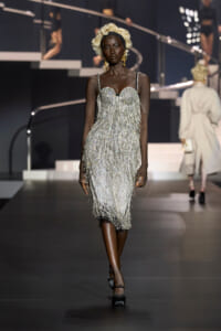 Black model walking a fashion runway in a silver beaded fringe dress, platinum curls and gold earrings.