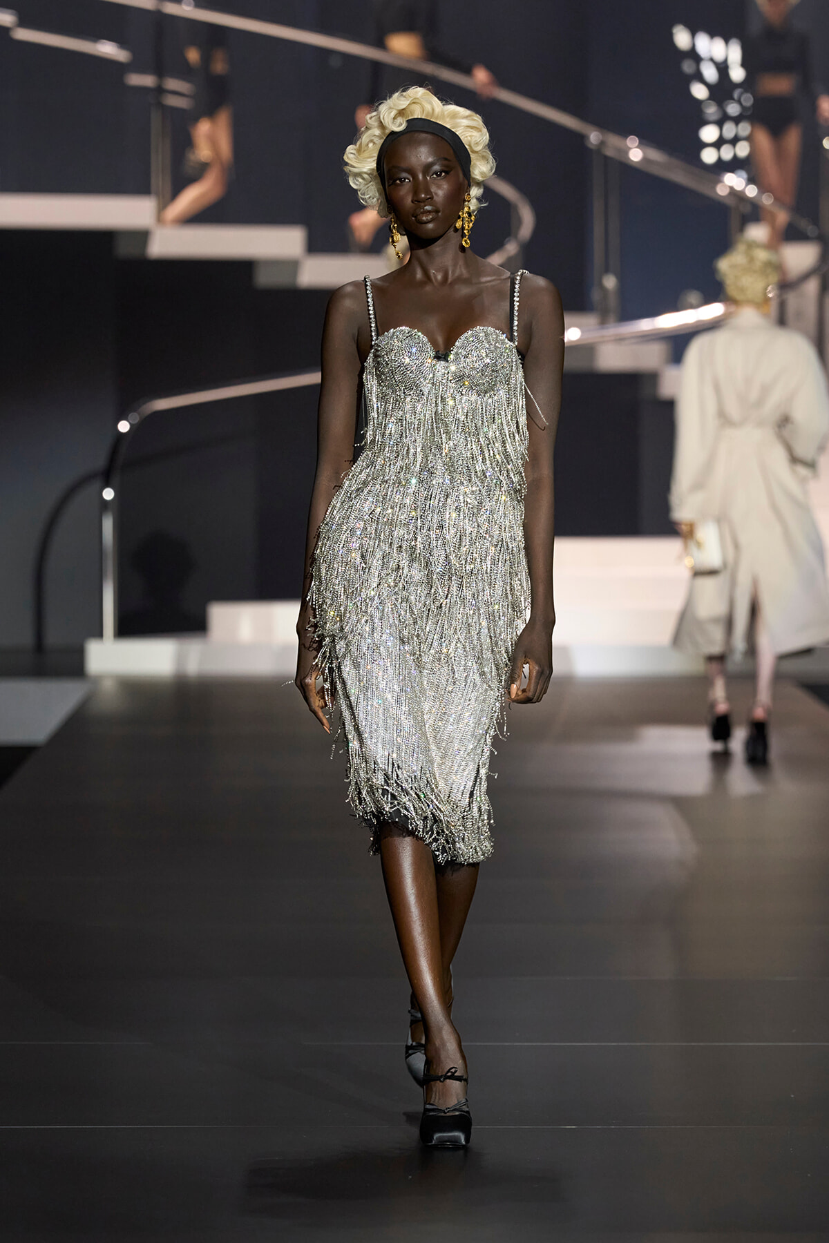 Black model walking a fashion runway in a silver beaded fringe dress, platinum curls and gold earrings.