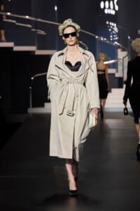 Female model in a beige belted trench coat with a black bra, oversized sunglasses, and gold earrings walking the runway on a fashion show set.