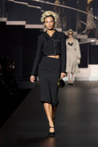 Model on a runway wearing a cropped black double-breasted jacket and a matching midi skirt, holding a small round black handbag.