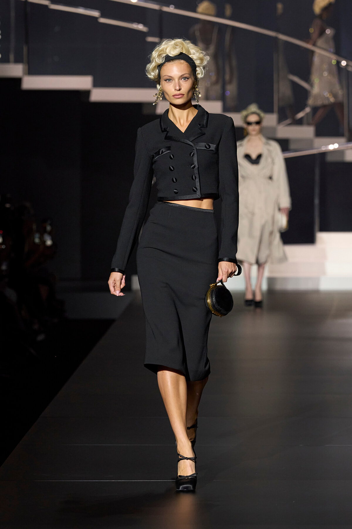 Model on a runway wearing a cropped black double-breasted jacket and a matching midi skirt, holding a small round black handbag.