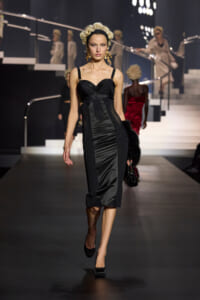 Model walking a runway in a black satin midi dress with a structured bodice and sheer panels, gold earrings.