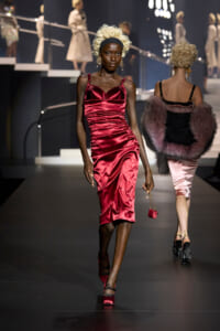 Model walks down the runway wearing a red satin slip dress with gathered bodice and black headband, blonde wig, and red heels." (informative)