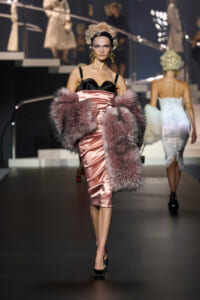 Model walking the runway in a pink satin dress with a black bustier, oversized pink fur stole, and black heels. Vintage curls and gold earrings complete the look.