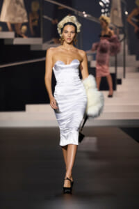 Model walks the runway in a fitted white satin strapless dress with a white fur stole draped over her arm.