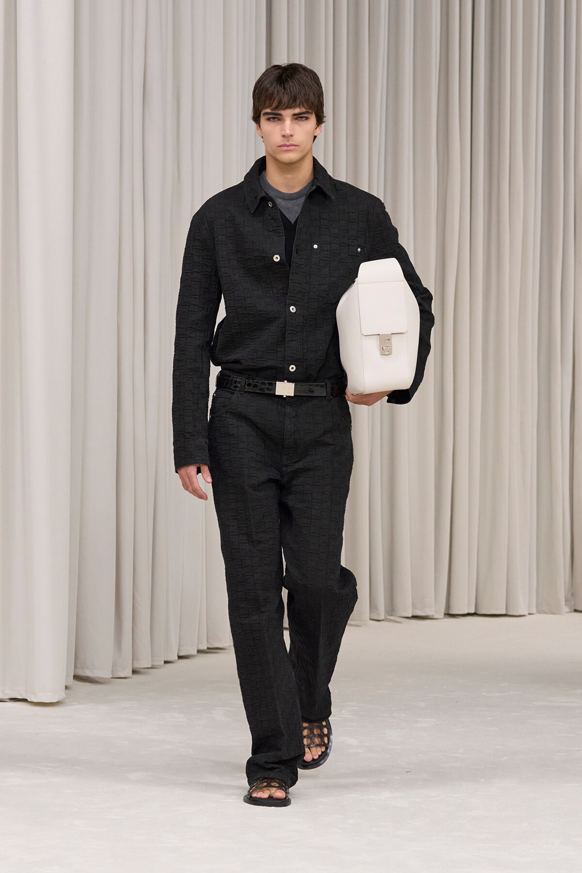 Male model walking on a runway in a black textured suit, holding a white rectangular bag under his arm.