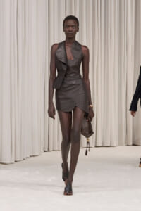 Fashion model walking a runway in a dark brown leather asymmetrical mini dress, with a textured brown clutch in hand against beige curtains.
