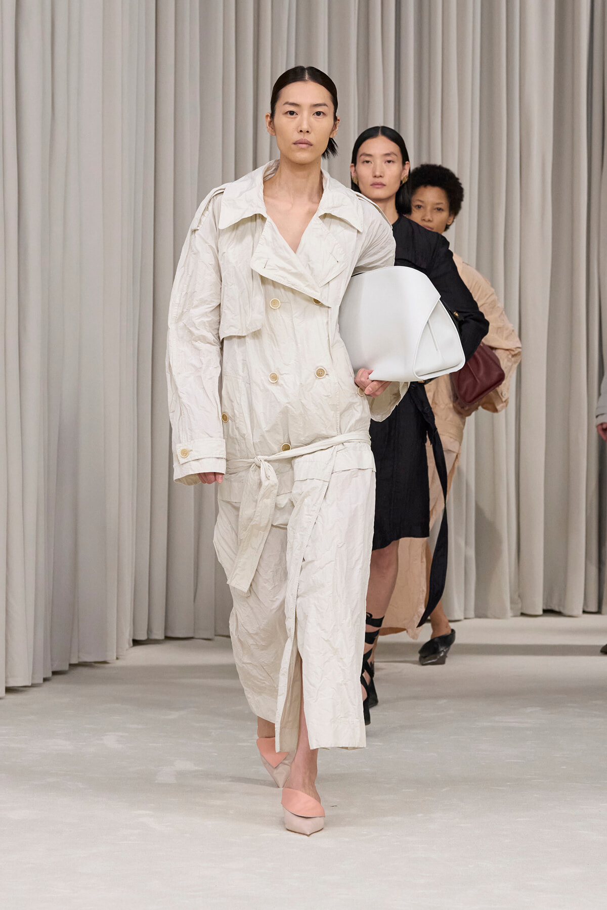 Model in a cream trench coat walking a runway, holding a white bag, wearing pink flats, with other models in neutral tones behind her.