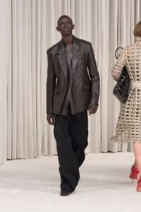 Male model in a dark leather blazer and loose black pants walking the runway.