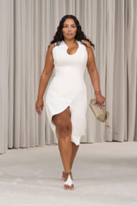 Model walks down the runway in a white asymmetrical dress, holding a beige clutch and wearing white sandals.