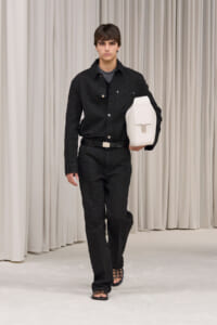 Male model in a black textured suit walking on a runway, carrying a white rectangular bag under his arm, beige curtain backdrop.