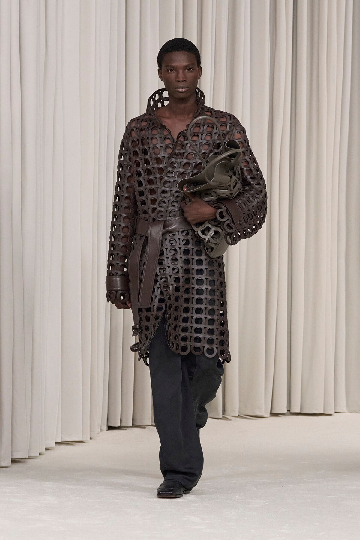 Model walks the runway in a brown circular-ring coat with a belt, carrying a matching bag.
