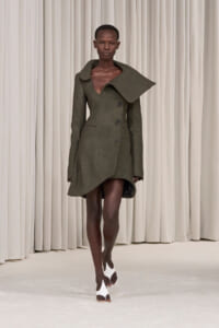 Model walking a runway in a textured olive-green coat with a large asymmetric collar and wrap-like hem, paired with white sandals.