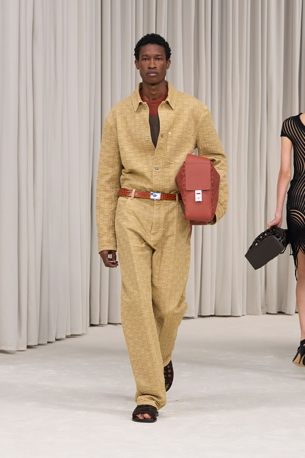 Male model walking a runway in a beige textured suit, brown belt, and brown sandals, carrying a red rectangular bag under his arm.