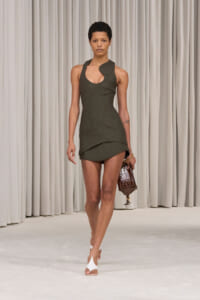 Model on a runway wearing a dark green asymmetrical mini dress, carrying a brown crocodile clutch under cream curtains.