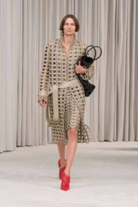 Model walks a runway in a beige circular-cutout dress with belt, carrying a black bag and wearing red heels.
