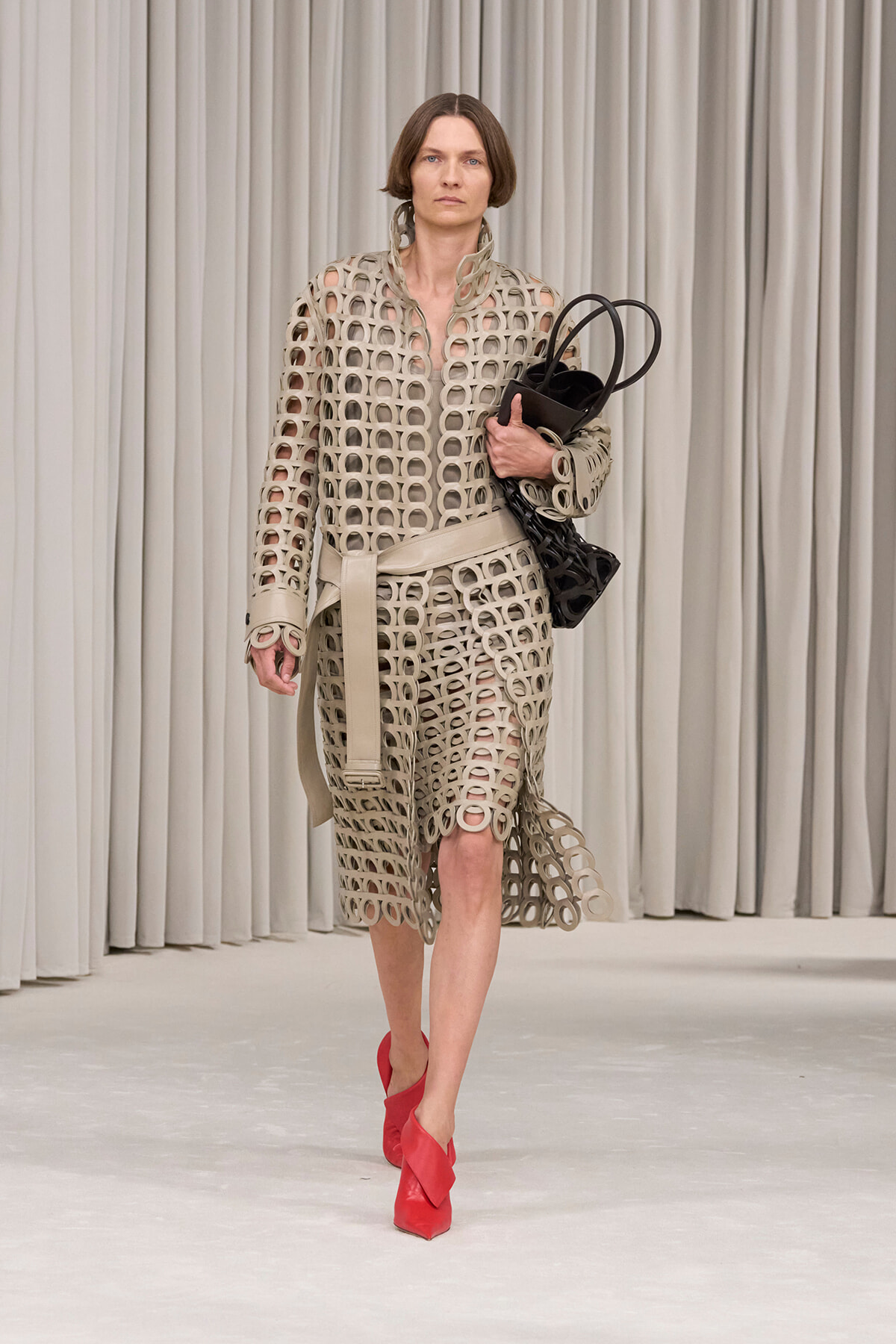 Model walks a runway in a beige circular-cutout dress with belt, carrying a black bag and wearing red heels.