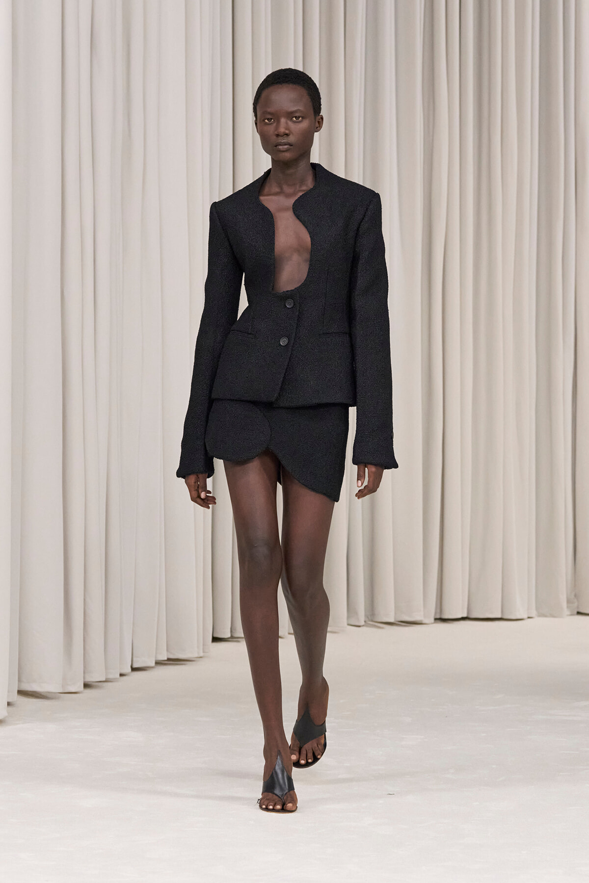 Runway model in a black tailored blazer with a curved-open front and matching mini shorts, wearing black sandals.
