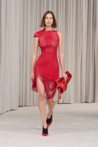 Model walking a runway in a red striped dress with fringe hem and matching red accessory in hand.