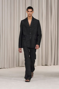 Male model walking a run-way in an oversized black suit with open neckline and wide-leg trousers, beige backdrop behind.