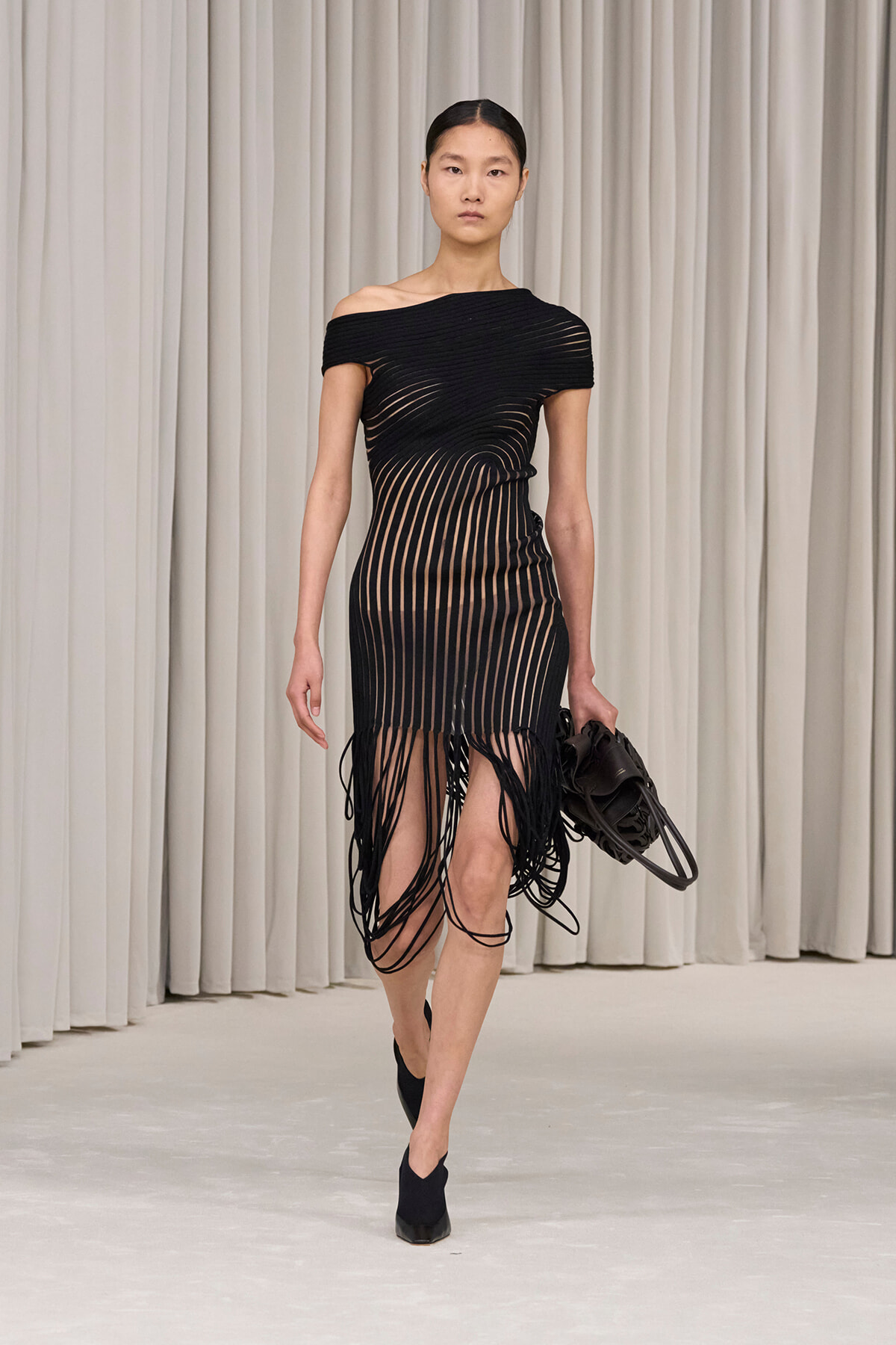 Model walks a runway in a black off-shoulder dress with long fringe strands, carrying a black handbag.