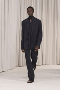 Male model in an oversized black suit with a high-neck scarf, walking on a white runway against light curtains.