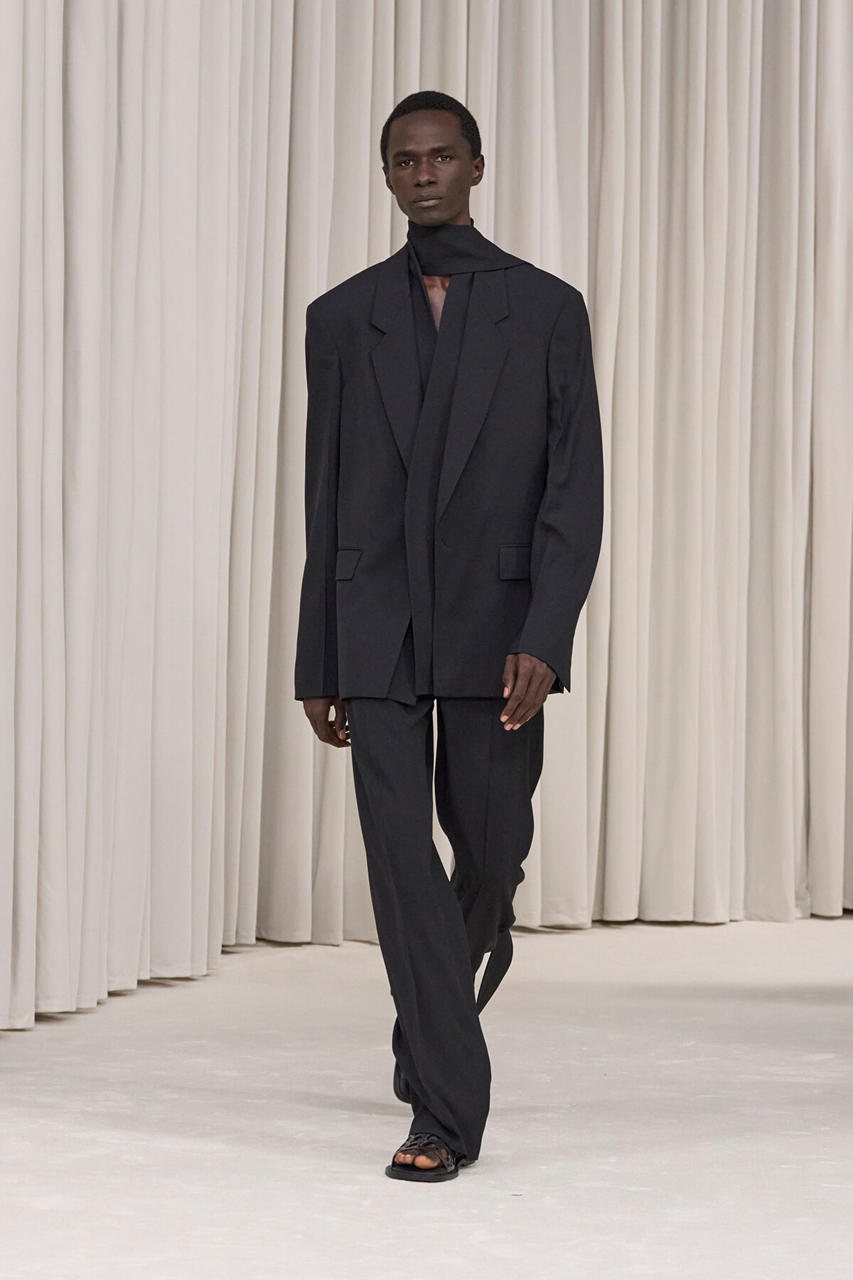 Male model in an oversized black suit with a high-neck scarf, walking on a white runway against light curtains.