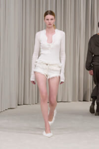 Model walking a fashion runway in a white ribbed top and frayed white shorts with furry slippers against a neutral backdrop.