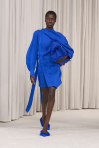 Model walking in a cobalt blue dress with voluminous sleeves, carrying a fringed matching bag and wearing fringed blue shoes on a white runway backdrop.