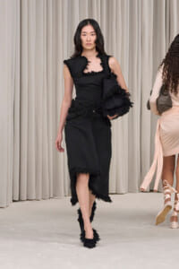 Model walking the runway in a black fringe-trim dress with a matching black bag.