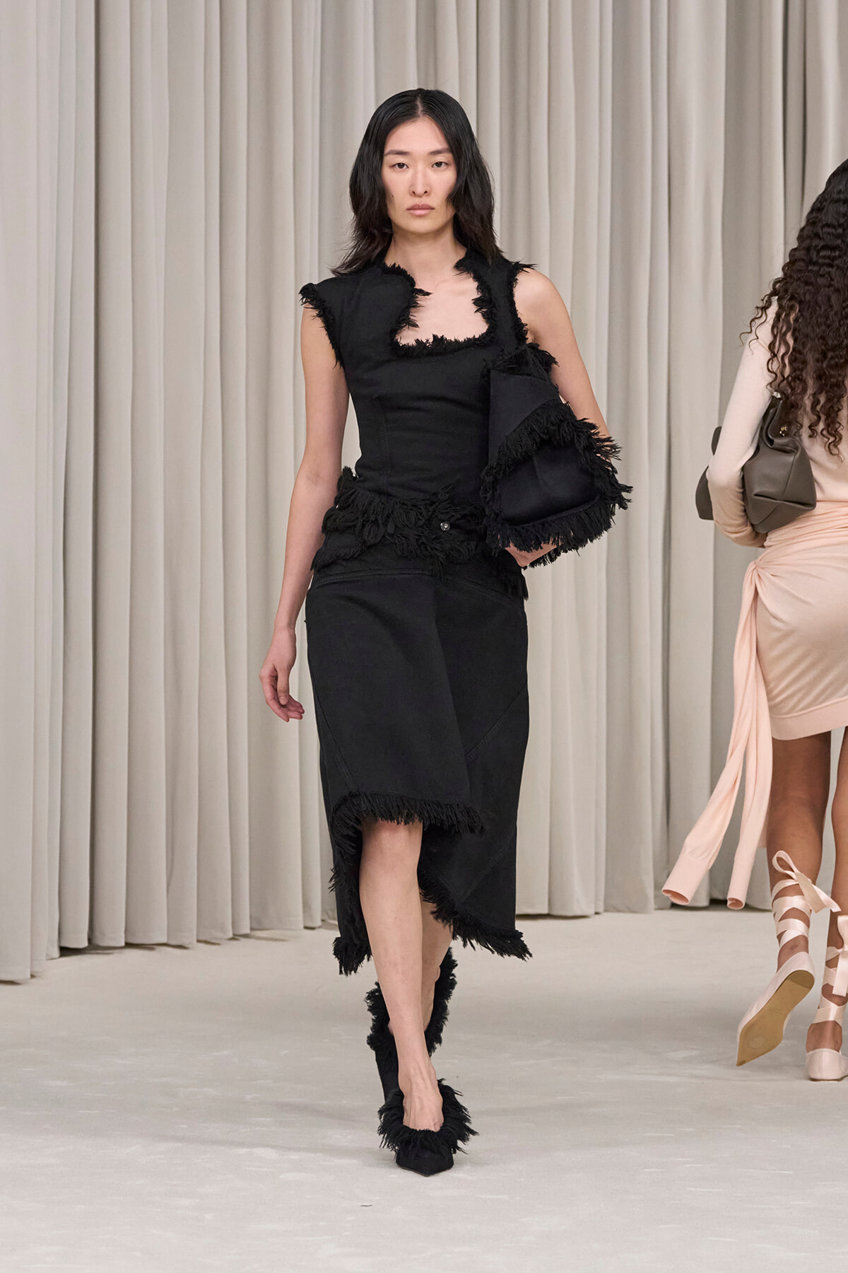 Model walking the runway in a black fringe-trim dress with a matching black bag.