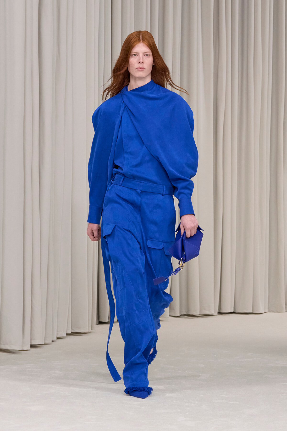 Model walks the runway in an all-blue draped top and cargo pants, holding a small blue handbag against a neutral backdrop.